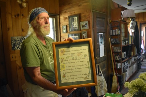 An older man with white hair and a beard holds a framed text in a eclectically decorated wooden room.